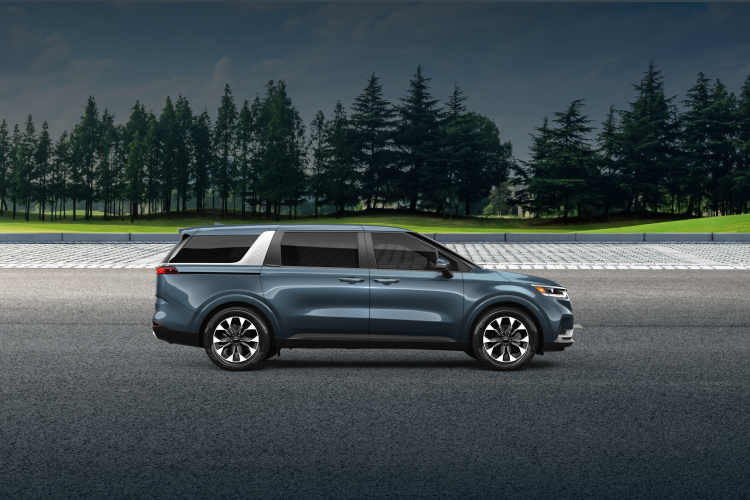 2024 Kia Carnival driving through an open road surrounded by grass fields and trees, side-view